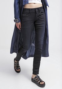 Svarta skinny jeans gjorda av stretchig denim, med klassiska fickor och en slät yta, i kombination med svarta remsandaler på en vit bakgrund.