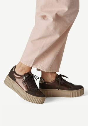 Brune metalliske platformsneakers med sorte snørebånd og sidelommer med lynlås, båret med lyserøde bukser mod en ensfarvet baggrund.