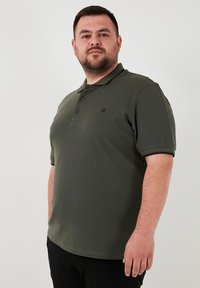 Zelený polo tričko vyrobené z mäkkej textílie, s trojzubkovou légou a malým logom na hrudi. Klasický golier a krátke rukávy.