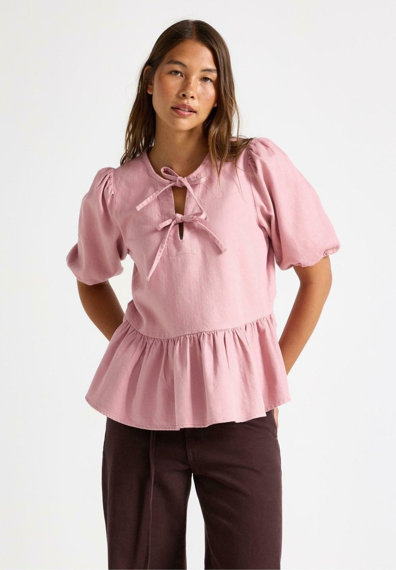 Lyserød bluse med pufærmer, en nøglehul-udskæring bundet med en sløjfe og en peplum-nederdel. Lavet af blødt, struktureret stof.