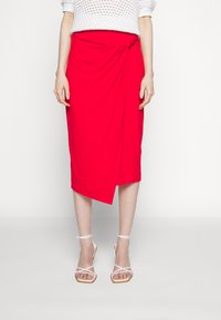 Rode knielange wikkelrok van zacht materiaal, met een geknoopte detail bij de taille. Gepaard met witte sandalen met straps.