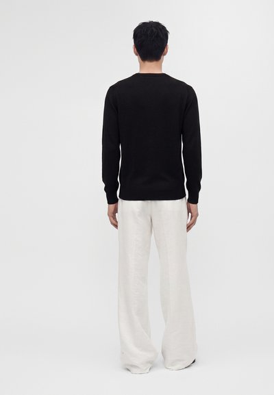 Homme de dos portant une chemise noire à manches longues et un pantalon blanc ample, se tenant devant un fond clair uni.