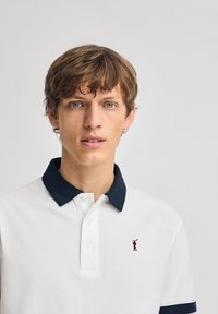 Hvid polo-shirt med en mørk navy krave, med en lille rød broderet golfspillerlogo. Bomuldsstof, tre-knaps placket, korte ærmer.