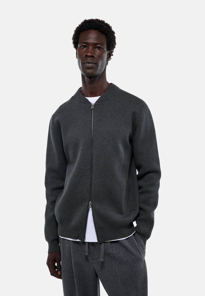 Sweat-shirt zippé gris foncé avec un col arrondi, des poches latérales et des poignets côtelés. Porté par-dessus une chemise blanche et associé à un pantalon gris.