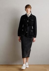 Giacca di jeans nera con doppie tasche sul petto, vita con cintura, abbinata a una gonna di jeans nera e scarpe da ginnastica bianche.