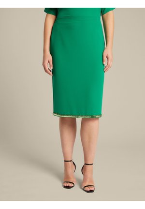 Groene knielange kokerrok met een gladde textuur, met een bewerkt zoomdetail van kraaltjes. Draagt met zwarte sandalen met hakken en bandjes.