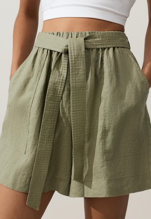 Pantaloncini di lino verde oliva a vita alta con ampia cintura di tessuto annodata davanti, tasche laterali, indossati con un top corto bianco.