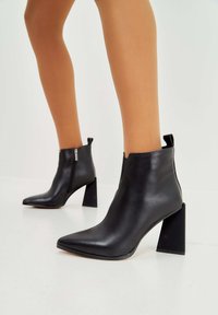 Botas de cuero negras hasta el tobillo con puntera afilada, cierre de cremallera lateral y un tacón en bloque grueso. Textura suave, diseño minimalista.