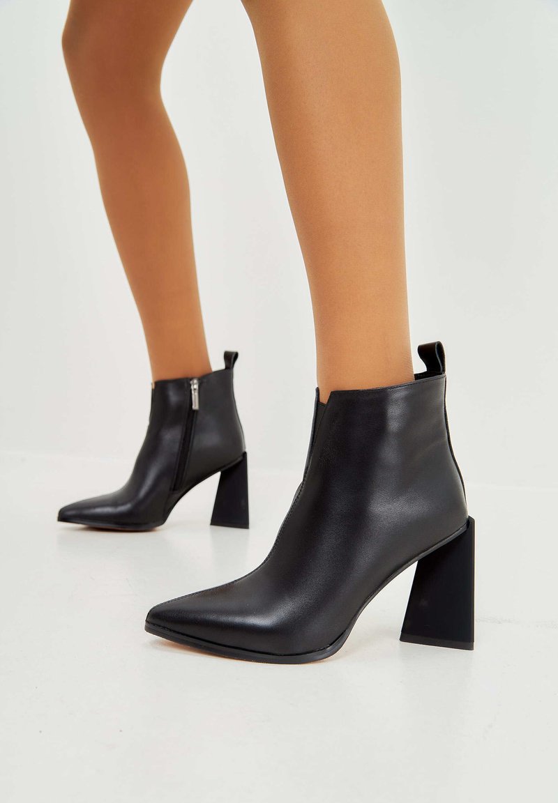 Botas de cuero negras hasta el tobillo con puntera afilada, cierre de cremallera lateral y un tacón en bloque grueso. Textura suave, diseño minimalista.