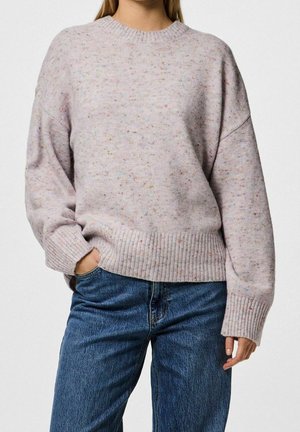 Woman wearing light pink speckled knit sweater with ribbed cuffs and hem, paired with blue denim jeans, one hand in pocket.