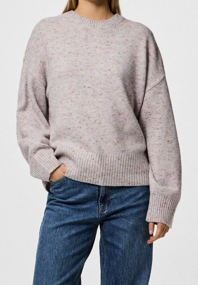 Mujer vistiendo un suéter de punto de color rosa claro con motas, con puños y dobladillo acanalados, combinado con jeans de mezclilla azules, con una mano en el bolsillo.