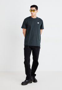 T-shirt di cotone verde scuro con una piccola toppa con logo, abbinata a jeans neri e stivali neri con cuciture gialle. Occhiali da sole trasparenti indossati.