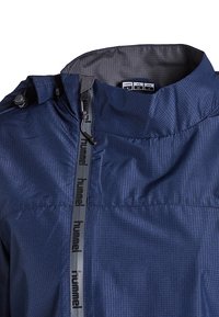 Giacca a vento blu navy con superficie testurizzata, colletto alto e zip verticale con un cinturino logo nero lungo la parte anteriore.