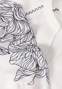 Tissu blanc avec une broderie florale noire détaillée et une couture en zigzag le long du bord, mettant en valeur un savoir-faire textile raffiné.