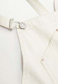 Gros plan sur la bretelle d'une salopette en denim couleur crème avec une boucle argentée, et une partie de la poche avant montrant des rivets en cuivre.