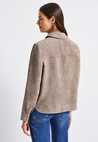 Frau mit langen braunen Haaren, die eine beigefarbene Cordjacke und blaue Jeans trägt, von hinten vor einem schlichten weißen Hintergrund gezeigt.