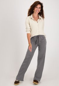 Vrouw die glimlacht, staand met één been naar voren gekruist, gekleed in een beige shirt met opgerolde mouwen, grijze wijde broek enbruine schoenen.