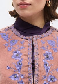 Κοντινό πλάνο ατόμου που φορά ροζ μπουφάν με μπορντούρα από λουλουδένια κεντήματα σε λεβαντί απόχρωση και διακοσμητική χάντρα πάνω σε σκούρο μωβ ζιβάγκο, με χρυσούς κρίκους σκουλαρικιών.