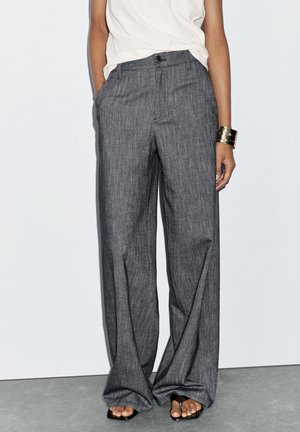 Personne portant un pantalon gris à jambes larges, un haut blanc, des sandales noires à bout carré et un large bracelet doré au poignet droit. Mains dans les poches.