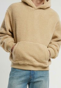Sudadera de forro polar beige con superficie texturizada, corte oversize, bolsillo tipo canguro y puños y dobladillo de canalé; llevada sobre unos vaqueros de mezclilla azules.