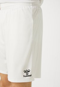 Witte sportshorts met een elastische tailleband en een zwart Hummel-logo nabij de onderzoom op het dijbeen van een persoon.