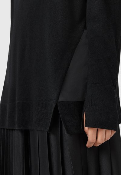 Pull en tricot noir avec fentes latérales et trous pour les pouces porté sur une jupe plissée noire, montrant un gros plan de la main et des détails du vêtement.