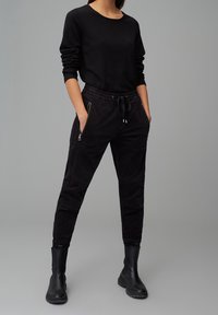 Zwart loungewear set bestaande uit een lange mouwen top en een broek met touwtjes en ritszakken. Gladde stof en aansluitende pasvorm.