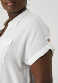 Bílá blůza s krátkým rukávem, s manžetou na knoflík a kapsou na hrudi. Hladká látka, design límečku a minimalistické detaily.