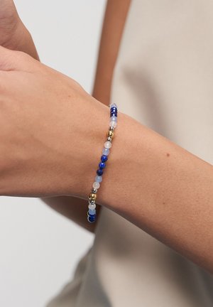 Armband mit abwechselnd blauen, weißen, silbernen und goldenen Perlen, getragen am Handgelenk einer Person vor neutralem Hintergrund.