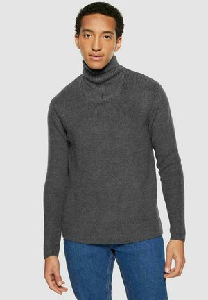 Jonge man die een donkergrijze geribbelde coltrui en blauwe jeans draagt, staand tegen een effen lichte achtergrond.