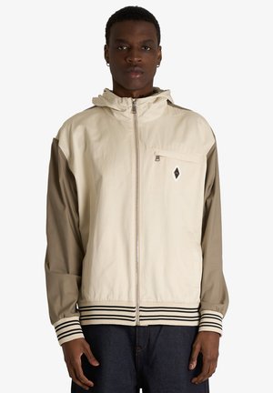 Modello maschile che indossa una giacca beige e oliva con zip, polsini e orlo a righe, in piedi davanti a uno sfondo bianco uniforme.