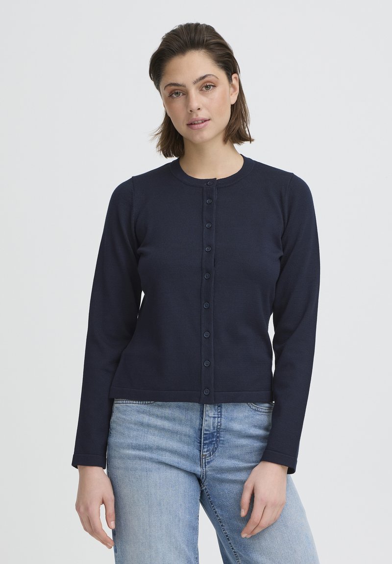 Cardigan bleu marine à manches longues en tissu côtelé, avec une fermeture boutonnée sur le devant et un col arrondi, porté avec un jean bleu clair.