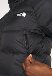 Giacca a vento nera con una tasca con zip, caratterizzata da un motivo cucito e il logo bianco "The North Face" sul petto.