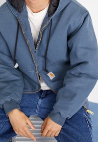 Blauwe rits-hoodie met een zwarte binnenvoering, elastische manchetten en een voorknopzak. Logo-etiket zichtbaar. Draag over een wit shirt en jeans.