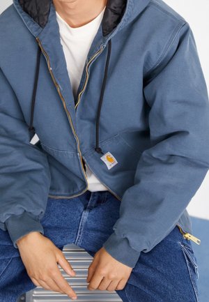 Blauwe rits-hoodie met een zwarte binnenvoering, elastische manchetten en een voorknopzak. Logo-etiket zichtbaar. Draag over een wit shirt en jeans.