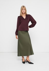 O bluză lungă burgundy cu decolteu în V, asociată cu o fustă midi din satin de culoare verde măsliniu. Ansamblul este completat de tocuri negre cu vârf ascuțit.