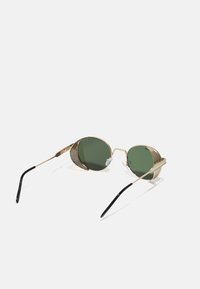 Lunettes de soleil rondes avec verres verts, monture en métal doré, accents en maille texturée sur les côtés et embouts en acétate noir sur les branches.