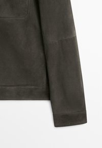 Dettaglio della manica e del fianco di una giacca in pelle scamosciata verde oliva scuro su sfondo bianco.