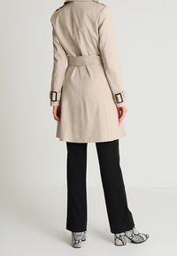 Beżowy płaszcz trencz z paskiem w talii, ozdobnymi naramiennikami i dopasowanym krojem, zestawiony z czarnymi spodniami i botkami w wężowy wzór.
