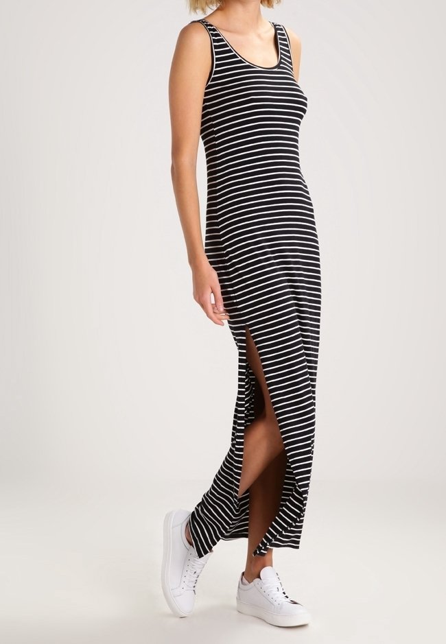 Femme portant une robe maxi sans manches rayée noir et blanc avec une fente latérale, associée à des baskets blanches, debout devant un fond uni.