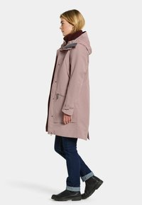 Chaqueta impermeable de color rosa claro con capucha, bolsillos frontales y un suave interior burdeos. Combinada con jeans oscuros y botines negros.