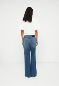 Wide-leg blue denim jeans with a high waist, five pockets, and a dark patch on the back waist; worn with a loose white t-shirt.