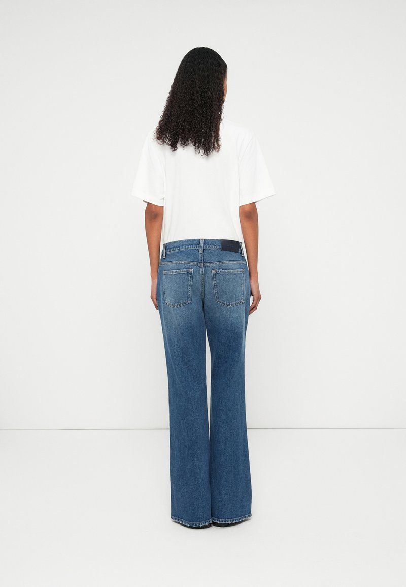 Jean en denim bleu à large jambe avec une taille haute, cinq poches et une pièce sombre à l'arrière de la taille ; porté avec un t-shirt blanc ample.