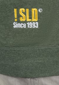 Sweat-shirt vert avec du texte brodé jaune et blanc : « ! SLD » et « Since 1993 ». Texture douce avec poignets et ourlet côtelés.