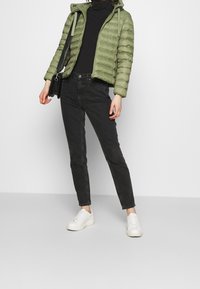 Grøn pufferjakke med lynlås, sort turtleneck, sorte slim jeans og hvide sneakers, sat mod en ensfarvet baggrund.