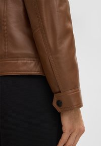 Mouwen van een bruin leren jack met gestikte details en een zwarte knop bij de manchet, gecombineerd met een gestructureerde zwarte stof eronder.