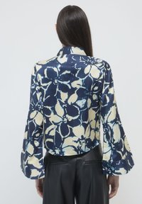 Femme portant un blouse à motifs floraux bleu marine et crème à manches longues, associée à un pantalon en cuir noir, vue de dos.