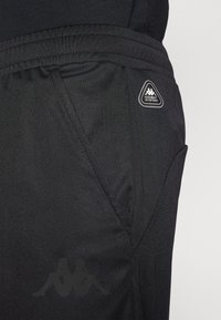 Zwarte sportbroek gemaakt van gladde stof. Voorzien van een elastische tailleband en een zichtbaar logo-opdruk aan de zijkant. Subtiele patroontextuur.