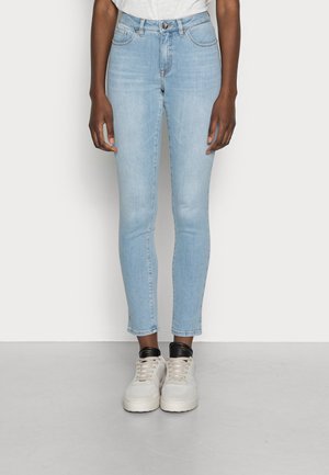 Person iført lyseblå slim-fit jeans og hvide sneakers med sorte detaljer, stående mod en ensfarvet lys baggrund.