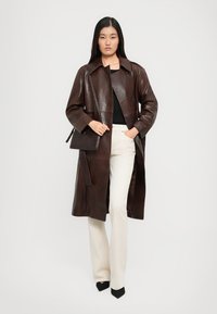 Manteau trench en cuir marron avec une taille ceinturée, de grands boutons et des poches latérales, porté sur un haut noir et un pantalon blanc.